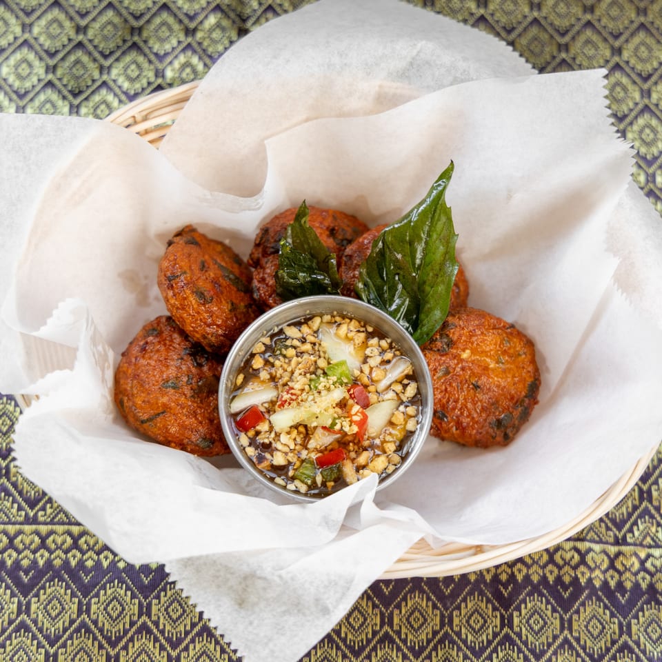 Fried fish cakes with a dipping sauce, garnished with basil, in a basket on a patterned tablecloth.