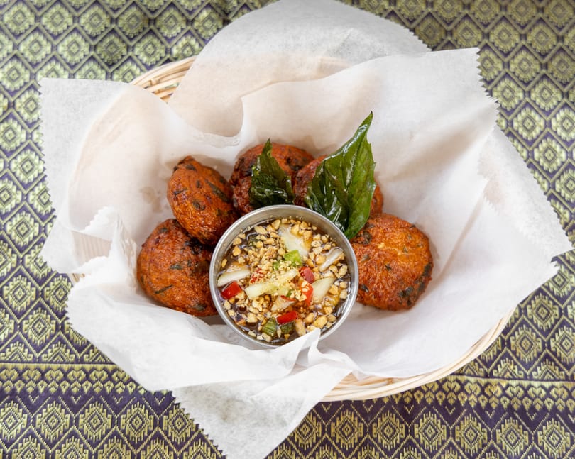 Fried fish cakes with a dipping sauce, garnished with basil, in a basket on a patterned tablecloth.