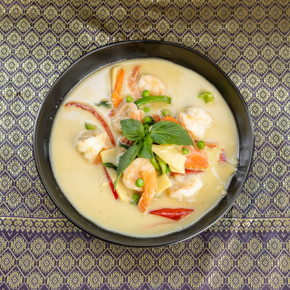 Creamy green curry with shrimp, bamboo shoots, peas, and basil in a black bowl on a patterned tablecloth.
