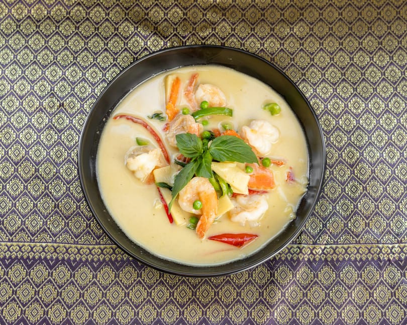 Creamy green curry with shrimp, bamboo shoots, peas, and basil in a black bowl on a patterned tablecloth.