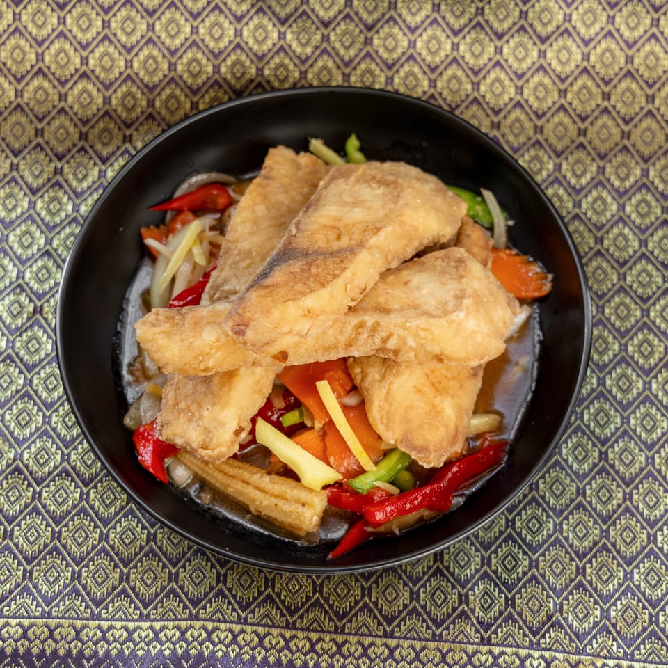 Crispy fried fish fillets with colorful vegetables in sauce, served in a black bowl on a patterned tablecloth.