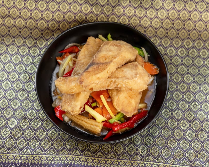 Crispy fried fish fillets with colorful vegetables in sauce, served in a black bowl on a patterned tablecloth.