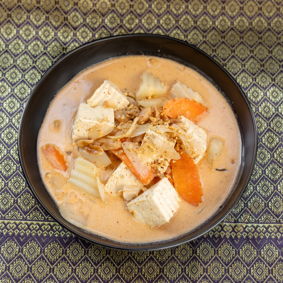 A black bowl of red curry with tofu, carrots, and onions on a purple and gold patterned tablecloth.