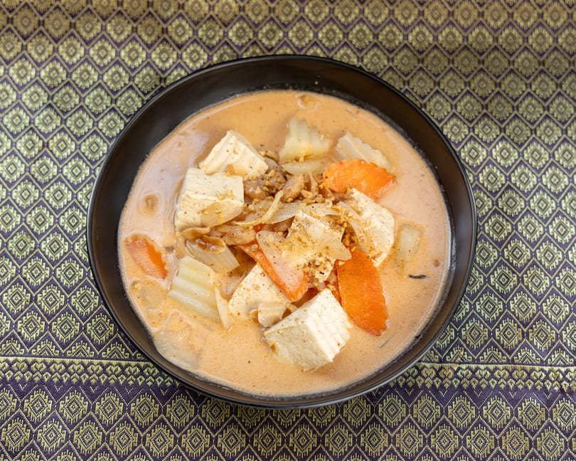 A black bowl of red curry with tofu, carrots, and onions on a purple and gold patterned tablecloth.