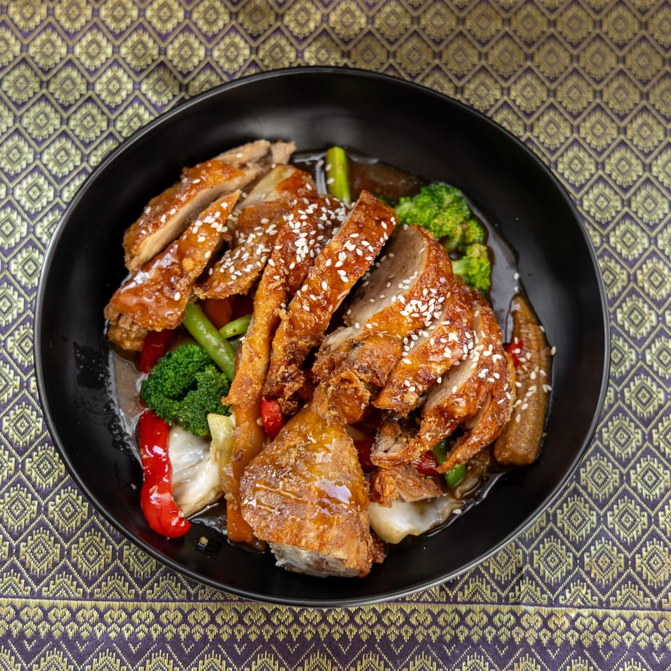 Crispy duck with sesame seeds, broccoli, and other vegetables in a black bowl on a patterned tablecloth.