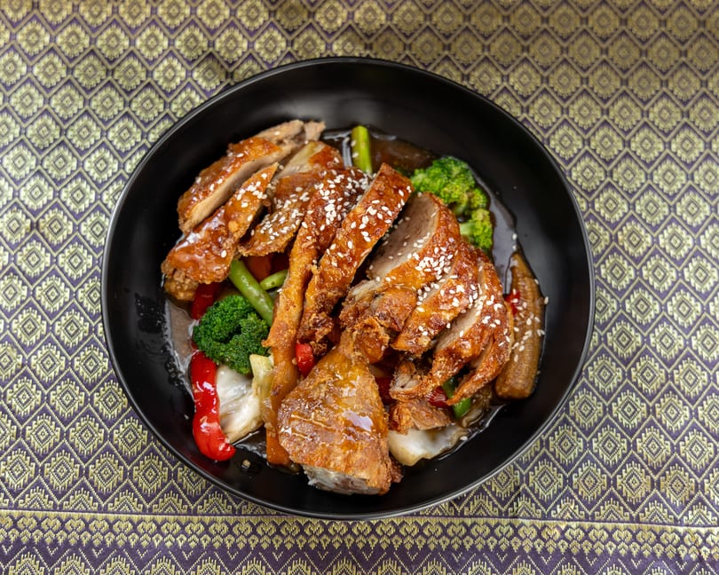 Crispy duck with sesame seeds, broccoli, and other vegetables in a black bowl on a patterned tablecloth.