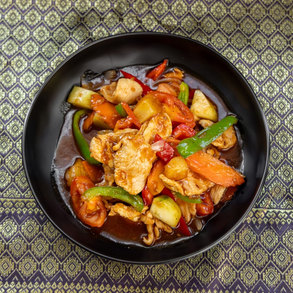 Sweet and sour chicken with pineapple, bell peppers, and carrots in a black bowl on a patterned tablecloth.