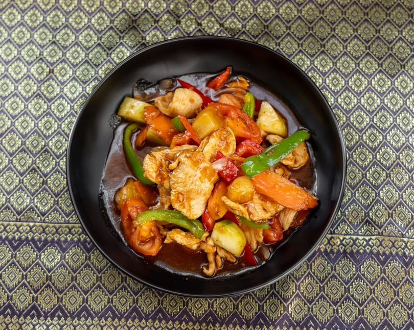 Sweet and sour chicken with pineapple, bell peppers, and carrots in a black bowl on a patterned tablecloth.