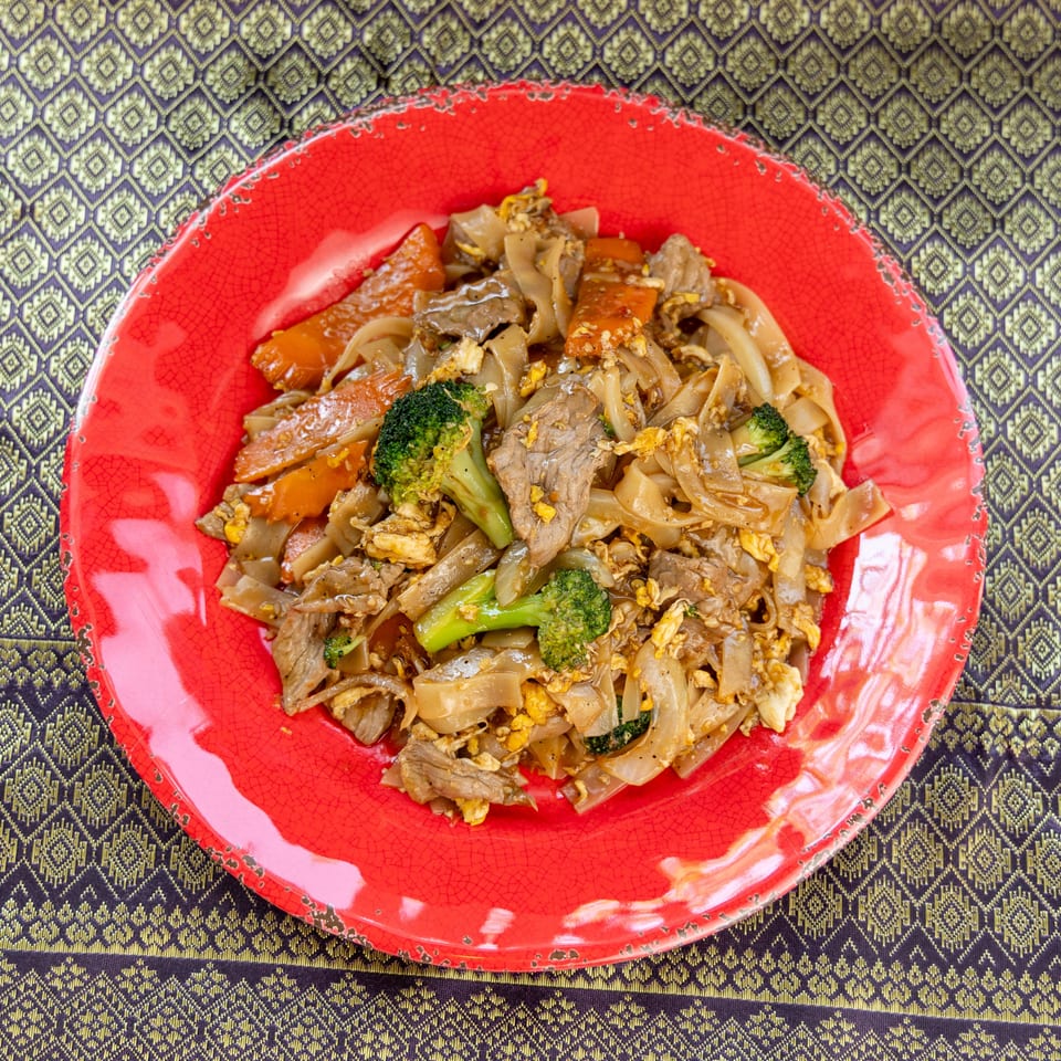 Pad See Ew with beef, broccoli, carrots, and egg on a red plate over a patterned tablecloth.