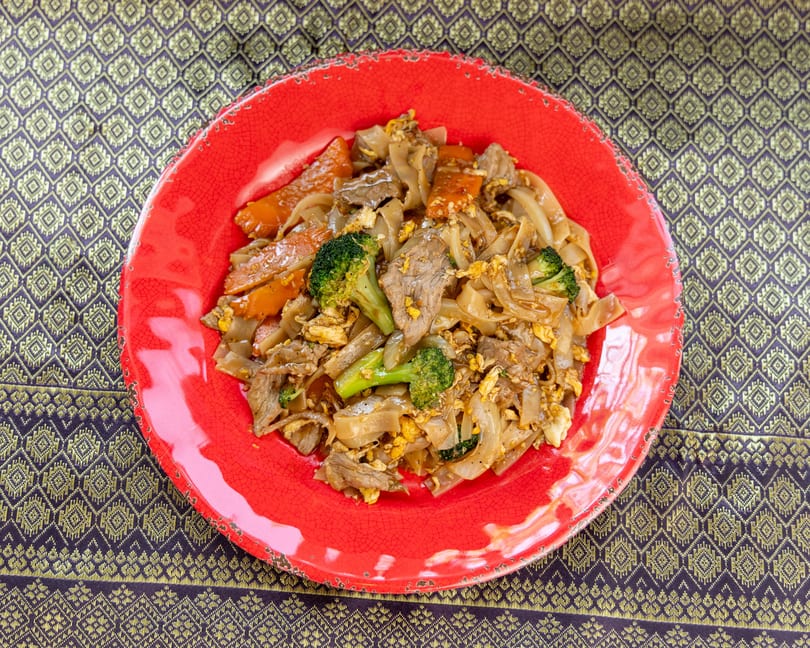 Pad See Ew with beef, broccoli, carrots, and egg on a red plate over a patterned tablecloth.