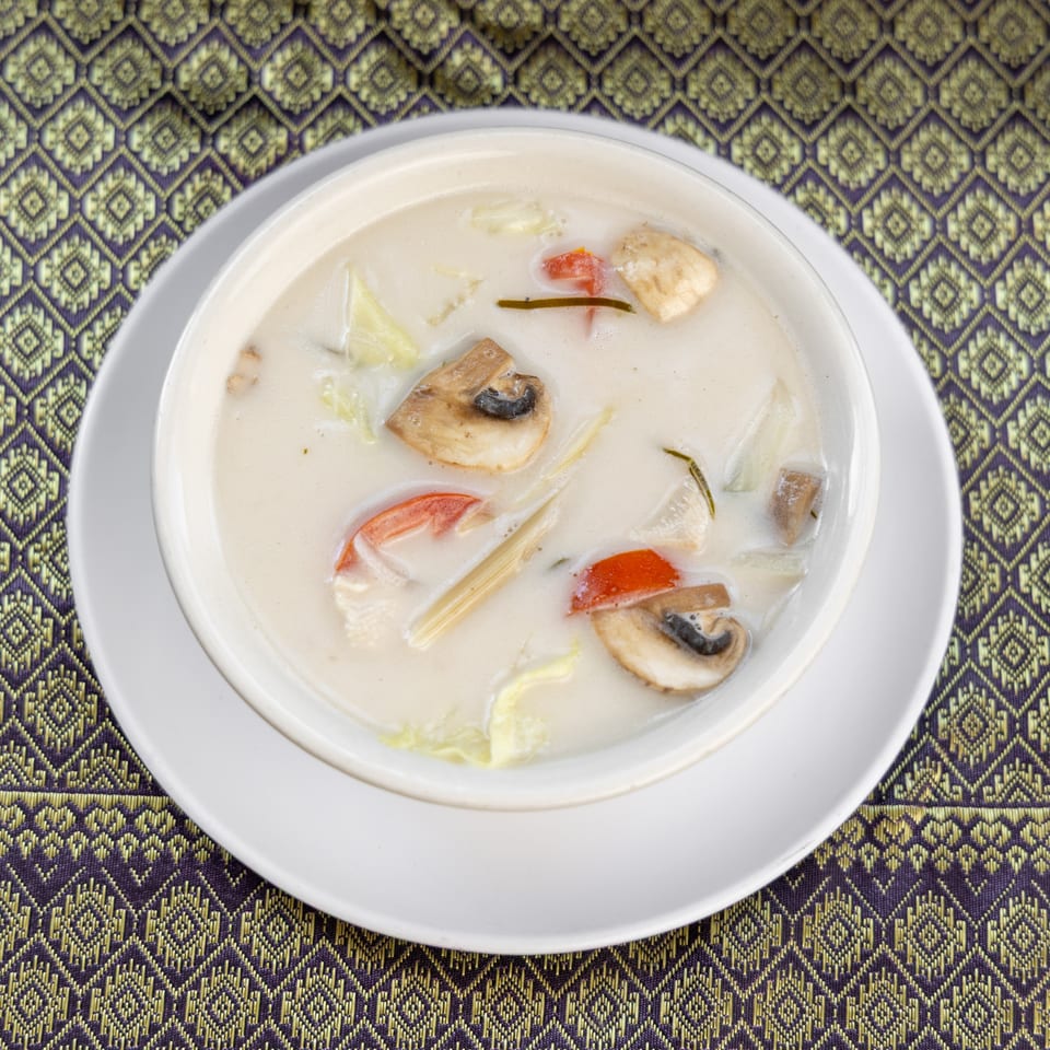 Creamy Tom Kha Gai soup with mushrooms, tomatoes, and lemongrass in a white bowl on a patterned mat.