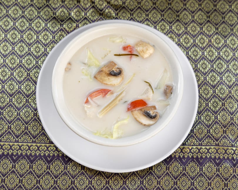 Creamy Tom Kha Gai soup with mushrooms, tomatoes, and lemongrass in a white bowl on a patterned mat.